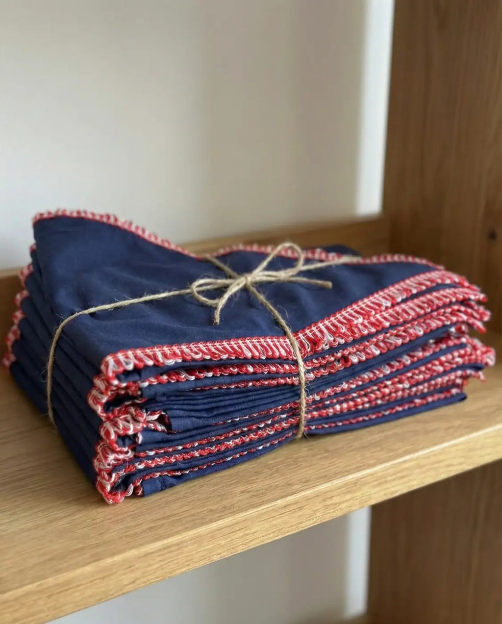 A stack of navy blue cor puro galão contorno polyester cloth napkins with red and white embroidered edges tied with twine l&l