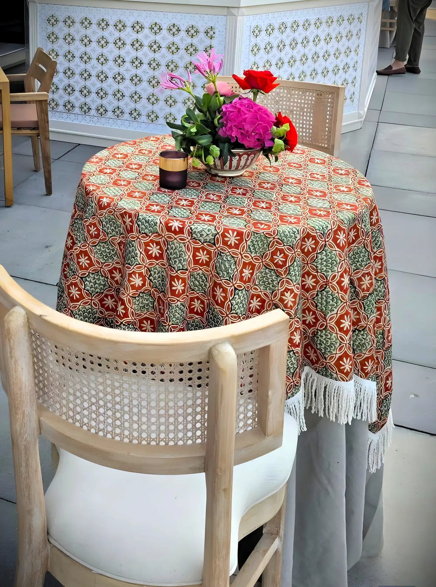 Uma toalha de mesa redonda ao ar livre com padrão vibrante de laranja e verde e franjas brancas, com drapeado sobre a mesa.